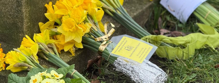 Several bouquets of yellow flowers and a small potted plant are placed at the base of a stone monument on grass.