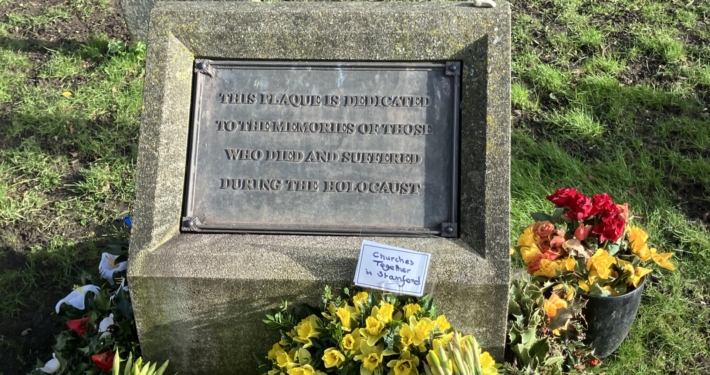 A memorial plaque dedicated to Holocaust victims is surrounded by yellow flowers and a small sign reading "Churches Together in Stratford.