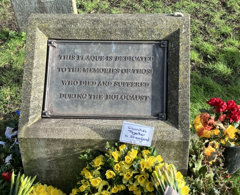 A memorial plaque dedicated to Holocaust victims is surrounded by yellow flowers and a small sign reading "Churches Together in Stratford.